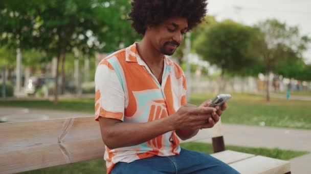 年轻的非洲裔美国人穿着衬衫坐在公园长椅上 用智能手机进行特写 微笑的男人在社交网络服务中通过手机进行交流 相机缩放 — 图库视频影像