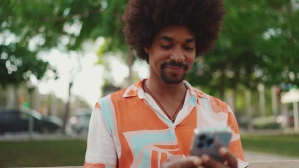 年轻的非洲裔美国人穿着衬衫坐在公园长椅上 使用智能手机的特写头像 微笑的男人在社交网络服务中的交流 相机在人的周围移动 — 图库视频影像