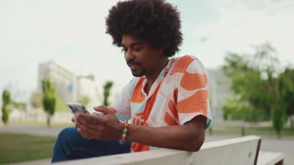 公園のベンチに座ってスマートフォンを使ってシャツを着ている若いアフリカ系アメリカ人の肖像画 ライフスタイルの概念 スローモーション — ストック動画