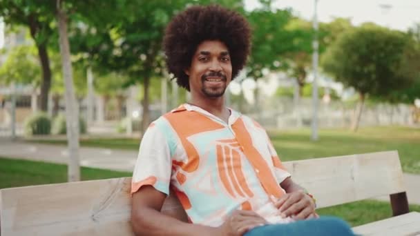 身穿衬衫 面带微笑的年轻非裔美国人坐在公园的长椅上 望着摄像机 这幅特写镜头让人感到轻松 生活方式的概念 慢动作 — 图库视频影像