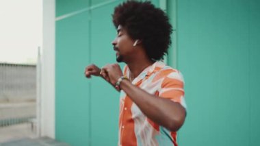 Tişört giyen, kablosuz kulaklıkla müzik dinleyen ve açık mavi duvar arkasında dans eden neşeli, genç Afrikalı Amerikalı adamın yakın çekimi. Yaşam tarzı konsepti. Yavaş çekim