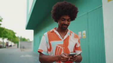 Sosyal ağlarda cep telefonuyla sohbet eden, açık mavi duvarlı arka planda yürüyen neşeli, Afro-Amerikalı genç bir adam. Yaşam tarzı konsepti. Yavaş çekim