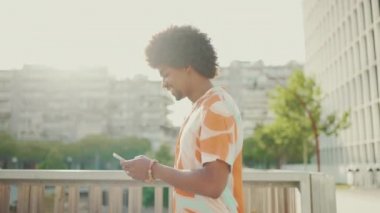 Tişörtlü, gülümseyen Afro-Amerikalı bir adamın yakın çekimi sosyal ağlarda cep telefonuyla güneşin altında yürürken yazıyor. Yaşam tarzı konsepti. Arka ışıklandırma. Yavaş çekim
