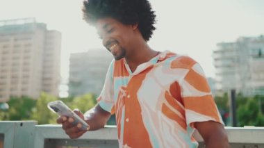Yakından bakınca, tişört giyen Afro-Amerikalı genç adam sosyal ağlarda şehir arka planında cep telefonuyla yazıyor. Kamera şahsa doğru hareket ediyor. Arka ışıklandırma. Yavaş çekim