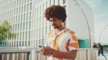 Gömlekli genç Afro-Amerikalı adamın yakın plan fotoğrafları sosyal ağlarda cep telefonuyla yazıyor ve şehir arka planında köprü boyunca yürüyor. Yaşam tarzı konsepti. Yavaş çekim