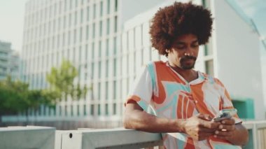 Yakından bakınca, tişört giyen Afro-Amerikalı genç adam sosyal ağlarda şehir arka planında cep telefonuyla yazıyor. Kamera şahsa doğru hareket ediyor. Kamera yakınlaştırılıyor. Yavaş çekim