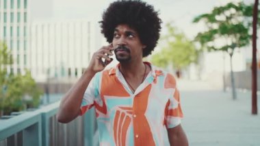 Gömlek giyen, cep telefonuyla konuşan ve şehir arka planında köprüde yürüyen Afrikalı genç bir adamın yakın çekimi. Yaşam tarzı konsepti. Yavaş çekim