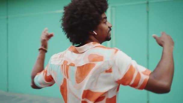 这位年轻快乐的非裔美国人穿着衬衫 用无线耳机听音乐 并在浅蓝色的墙壁背景下跳舞 生活方式的概念 慢动作 — 图库视频影像