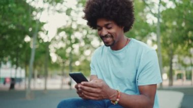 Açık mavi tişörtlü, şehir meydanındaki bankta oturan ve akıllı telefonunu kullanan Afrikalı genç bir adam. Sosyal ağ hizmetlerinde iletişim kuran adam. Yaşam tarzı konsepti. yavaş çekim