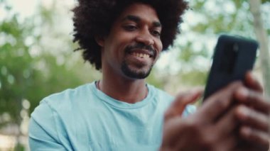 Açık mavi tişörtlü, şehir bankında oturan ve akıllı telefonunu kullanan Afro-Amerikan bir adamın yakın plan portresi. Adam fotoğraflara, cep telefonundaki videolara bakıyor. Yaşam tarzı konsepti. yavaş çekim