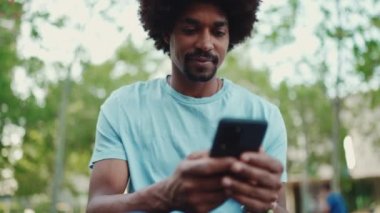 Açık mavi tişörtlü Afro-Amerikalı genç adamın şehir bankında oturduğu fotoğraflara ve cep telefonundaki videolara yakından baktığı fotoğraf. Kamera ileri doğru hareket ediyor. yavaş çekim