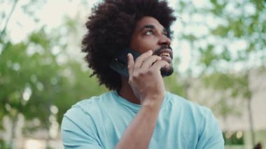 Yakından bakınca açık mavi tişörtlü Afro-Amerikan bir adam şehir bankında oturuyor ve akıllı telefondan konuşuyor. Yaşam tarzı konsepti. yavaş çekim
