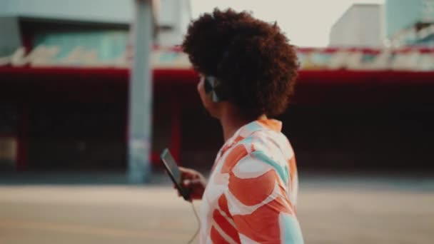 ヘッドフォンで音楽を聴いたり 都会の街の背景で踊ったりするシャツを着た陽気な若いアフリカ系アメリカ人の男性のクローズアップ ライフスタイルの概念 スローモーション — ストック動画