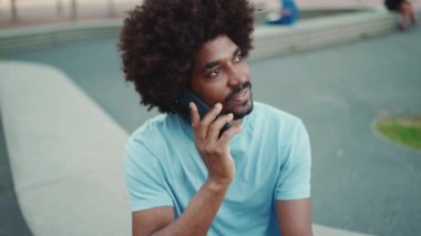 Yakından bakınca açık mavi tişörtlü Afro-Amerikan bir adam şehir bankında oturuyor ve akıllı telefondan konuşuyor. Kamera şahsa doğru hareket ediyor. Yaşam tarzı konsepti. yavaş çekim