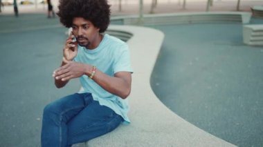 Açık mavi tişörtlü genç Afro-Amerikalı adam şehir bankında oturuyor ve akıllı telefondan konuşuyor. Yaşam tarzı konsepti. yavaş çekim