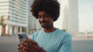 Açık mavi tişörtlü genç Afro-Amerikan bir adamın akıllı telefonunu kullanarak yakın plan portresi. Adam fotoğraflara, cep telefonundaki videolara bakıyor. yavaş çekim