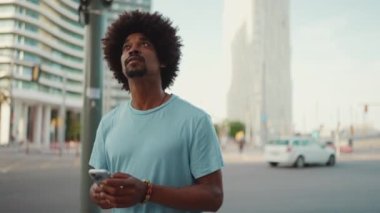 Yakından bakınca, açık mavi tişörtlü Afro-Amerikan bir adam elinde akıllı telefonuyla modern şehrin sokaklarında geziyor ve telefon ekranında elektronik haritalı bir adres arıyor. yavaş çekim