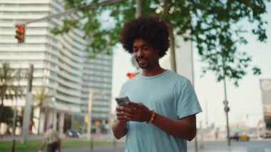 Yakından bakınca, açık mavi tişörtlü Afro-Amerikan bir adam elinde akıllı telefonuyla modern şehrin sokaklarında geziyor ve telefon ekranında elektronik haritalı bir adres arıyor. yavaş çekim