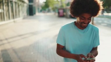 Açık mavi tişörtlü, akıllı telefonunu kullanan Afro-Amerikan genç adamın yakın çekimi. Adam fotoğraflara, cep telefonundaki videolara bakıyor. Arka ışık. Yavaş çekim