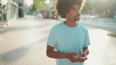 Açık mavi tişörtlü genç Afro-Amerikan bir adam sokakta yürürken ve akıllı telefonunu kullanırken. Adam fotoğraflara, cep telefonundaki videolara bakıyor. Arka ışık. Yavaş çekim