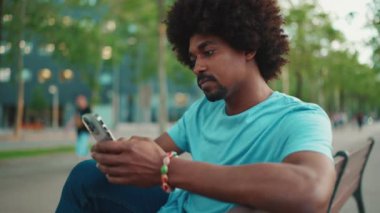 Yakından bakınca mavi tişörtlü Afro-Amerikan bir adam akıllı telefonunu kullanarak parktaki bankta oturuyor. Sosyal ağ hizmetlerinde iletişim kuran adam. Yaşam tarzı konsepti. yavaş çekim