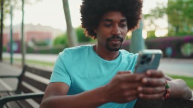 Yakından bakınca mavi tişörtlü Afro-Amerikan bir adam akıllı telefonunu kullanarak parktaki bankta oturuyor. Sosyal ağ hizmetlerinde iletişim kuran adam. Yaşam tarzı konsepti. yavaş çekim
