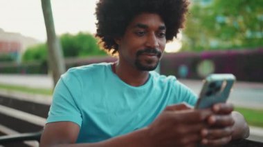 Yakından bakınca mavi tişörtlü Afro-Amerikan bir adam akıllı telefonunu kullanarak parktaki bankta oturuyor. Sosyal ağ hizmetlerinde iletişim kuran adam. Yaşam tarzı konsepti. yavaş çekim
