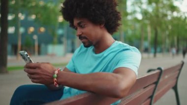 Yakından bakınca mavi tişörtlü Afro-Amerikan bir adam akıllı telefonunu kullanarak parktaki bankta oturuyor. Sosyal ağ hizmetlerinde iletişim kuran adam. Yaşam tarzı konsepti. yavaş çekim