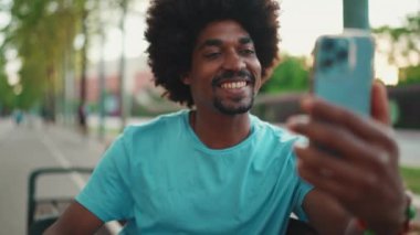 Mavi tişörtlü, parktaki bankta oturan ve hoparlörden konuşan Afro-Amerikan genç adamın yakın çekimi. Gülümseyen adam cep telefonundan sesli mesaj gönderiyor. Yaşam tarzı konsepti. Yavaş çekim