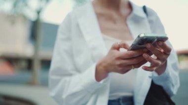 Genç bir kadının yakın çekimi akıllı telefonu elinde tutuyor ve haber yayınlarına aktarıyor. Kız elinin yakın çekimi şehir planında açık havada cep telefonu kullanıyor