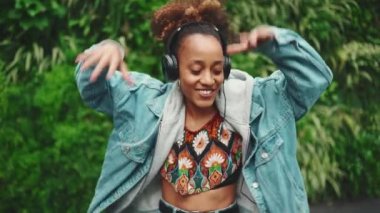 Kot ceketli, at kuyruklu gülümseyen Afrikalı kız şehir parkında yürüyor kulaklıkla müzik dinliyor ve yeşil ağaçların arkasında dans ediyor. Yavaş çekim