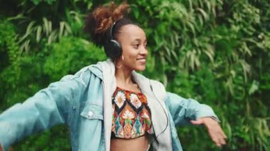 Kot ceketli, at kuyruklu gülümseyen Afrikalı kız şehir parkında yürüyor kulaklıkla müzik dinliyor ve yeşil ağaçların arkasında dans ediyor. Yavaş çekim