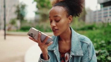 At kuyruklu, kot ceketli Afrikalı bir kızın yakın plan portresi cep telefonuna modern şehir arka planına sesli mesaj bırakıyor. Yavaş çekim