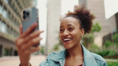 Kapanış portresi at kuyruklu, kot ceketli şirin Afrikalı kız, sabah kahvesiyle oturur ve modern şehir arka planında video görüşmesi yapar. Yavaş çekim