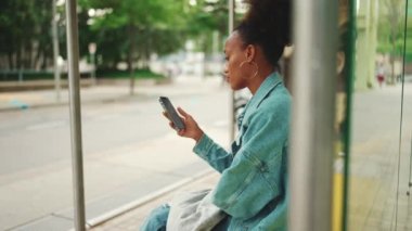 At kuyruklu, kot ceket giyen, ulusal desenli taraklı şirin Afrikalı kız otobüs durağında oturuyor ve cep telefonunu kullanıyor. Yavaş çekim