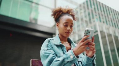 Kot ceketli şirin Afrikalı kız oturur ve fotoğraflara bakar, modern binaların arka planındaki cep telefonlarında videolar. Yavaş çekim
