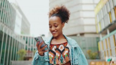 At kuyruklu şirin Afrikalı kız, kot ceket giyiyor, ulusal desenli tarlada, elinde telefonla modern binaların arka planına doğru yürüyor. Yavaş çekim
