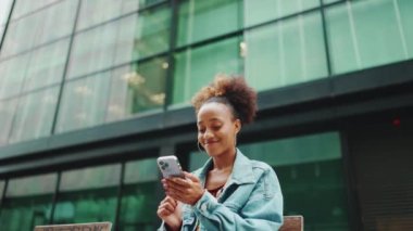 At kuyruklu, kot ceket giyen, milli desenli, bankta oturan ve modern binaların arka planında akıllı telefon kullanan tatlı Afrikalı kız. Yavaş çekim