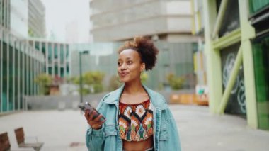 At kuyruklu şirin Afrikalı kız, kot ceket giyiyor, ulusal desenli tarlada, elinde telefonla modern binaların arka planına doğru yürüyor. Yavaş çekim