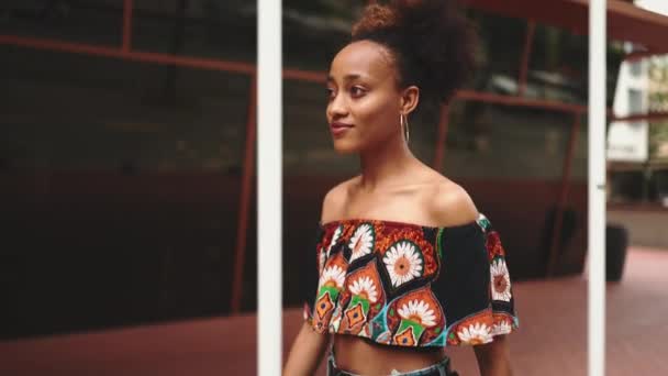 ポニーテールを国のパターンで作物の上に身に着けているとクローズアップアフリカの女の子は 近代的な建物の背景に対して通りを笑顔歩いています スローモーション — ストック動画