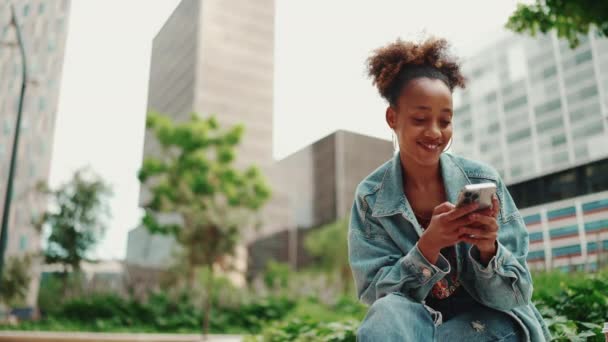 デニムジャケットを着たポニーテールを着たかわいいアフリカの女の子が座って 現代の建物の背景に携帯電話上の写真 ビデオを通して見ています スローモーション — ストック動画