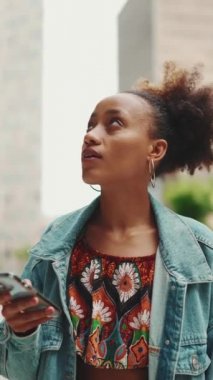 Modern şehrin caddesinde kot ceket giyen ve elinde akıllı telefon tutan yakın plan Afrikalı kız telefon ekranında şehirdeki elektronik haritalı adresi arıyor. Kamera yakınlaştırılıyor