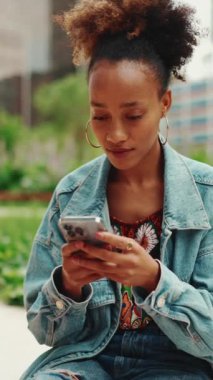 At kuyruklu şirin Afrikalı kız, kot ceket giyiyor, oturuyor ve fotoğraflara bakıyor, modern binaların arka planındaki cep telefonlarında videolar var. Yavaş çekim