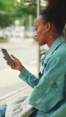 Yakından bakınca, at kuyruklu, kot ceketli, ulusal desenli tarlada, otobüs durağında oturan ve cep telefonunu kullanan tatlı Afrikalı bir kız. Yavaş çekim