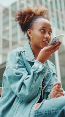 At kuyruklu, kot ceketli Afrikalı bir kızın yakın plan portresi modern şehir arka planında cep telefonuna sesli mesaj bırakıyor. Yavaş çekim