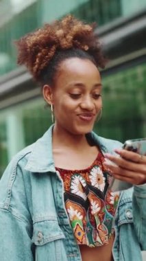 At kuyruklu, kot ceketli, ulusal desenli ekin giymiş, elinde telefonla modern binaların arka planına bakan sevimli Afrikalı kız.