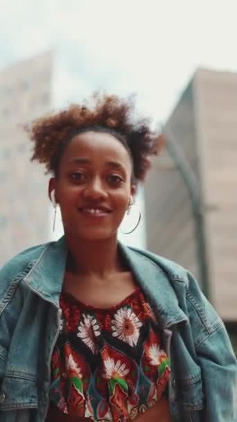 Verical Video Closeup ポニーテールのデニムジャケットを着た笑顔のアフリカの女の子 ヘッドフォンで音楽を聞いて屋外で踊る全国パターンのクロップトップ スローモーション — ストック動画