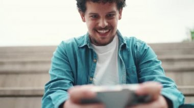 Yakından gülümseyen, kot gömlekli, yüksek basamaklarda oturan ve cep telefonu kullanan sakallı bir adam. Akıllı telefonda oynayan adam.