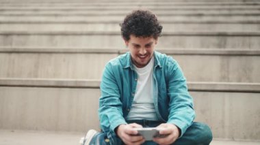 Kot gömlekli gülümseyen genç sakallı adam yüksek basamaklarda oturuyor ve cep telefonu kullanıyor. Akıllı telefonda oynayan adam.