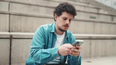 Yakından, kot gömlekli, gülümseyen sakallı genç adam yüksek basamaklarda oturuyor ve cep telefonu kullanıyor. Akıllı telefondan mesaj yazan adam